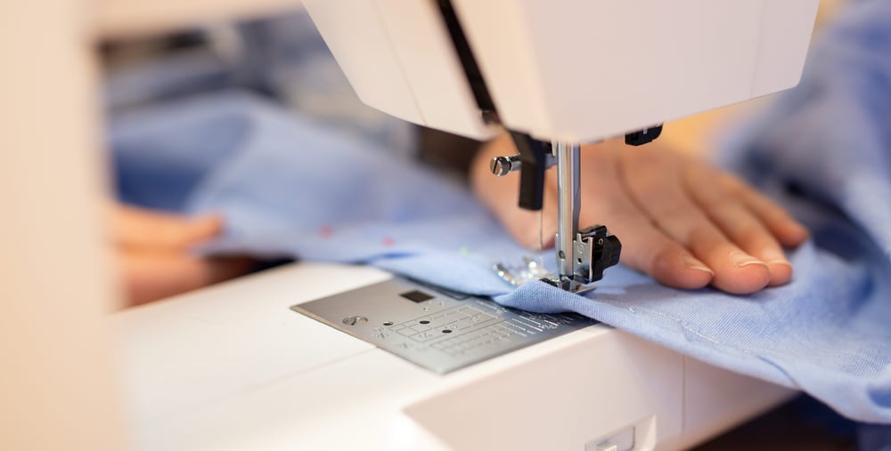 Närbild på en symaskin där en hand styr ett blått tyg genom nålen under sömnad.