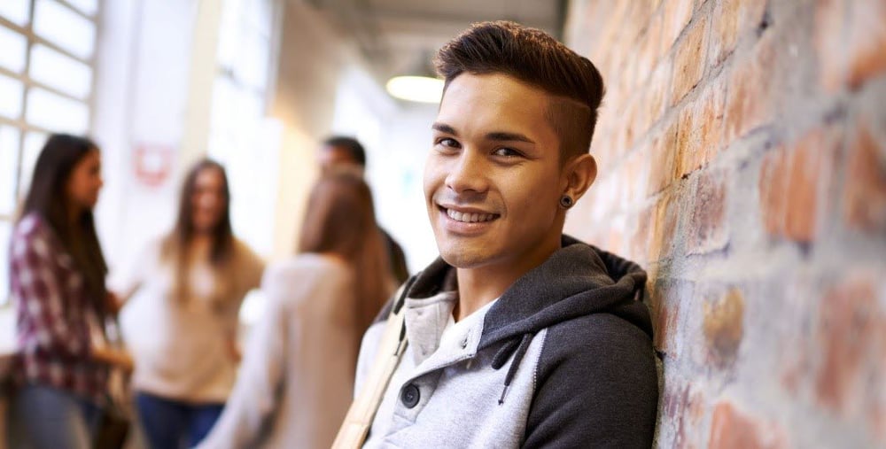 Ung person som står mot en tegelvägg, fyra personer står i bakgrunden och pratar med varandra. 