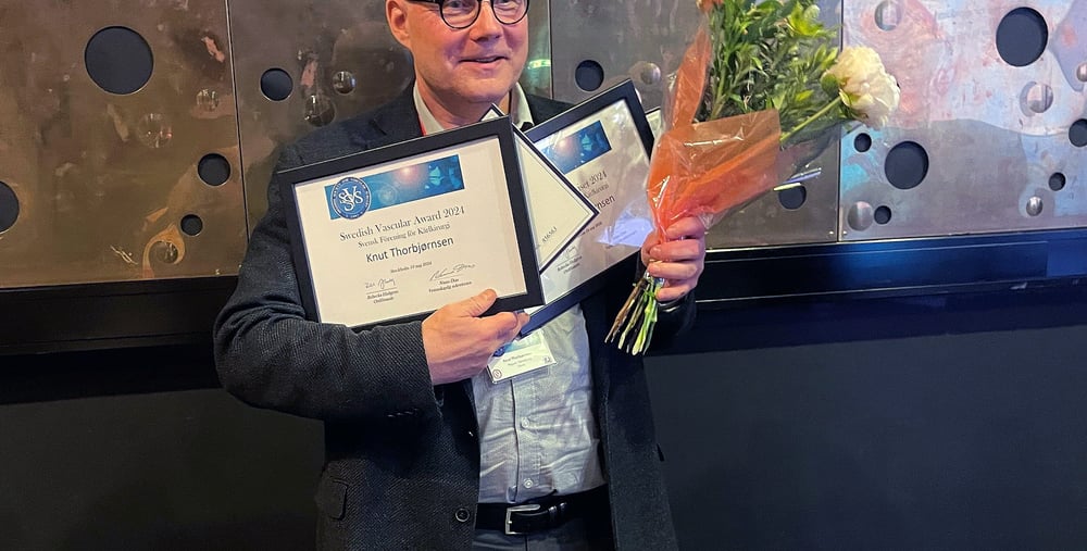 Kärlkirurgen Knut Thorbjørnsen, medelålders man står med blommor i handen och flera inramade prisdokument i handen. Han ler mot kameran. Han bär en ljus skjorta och en grå kavaj. 