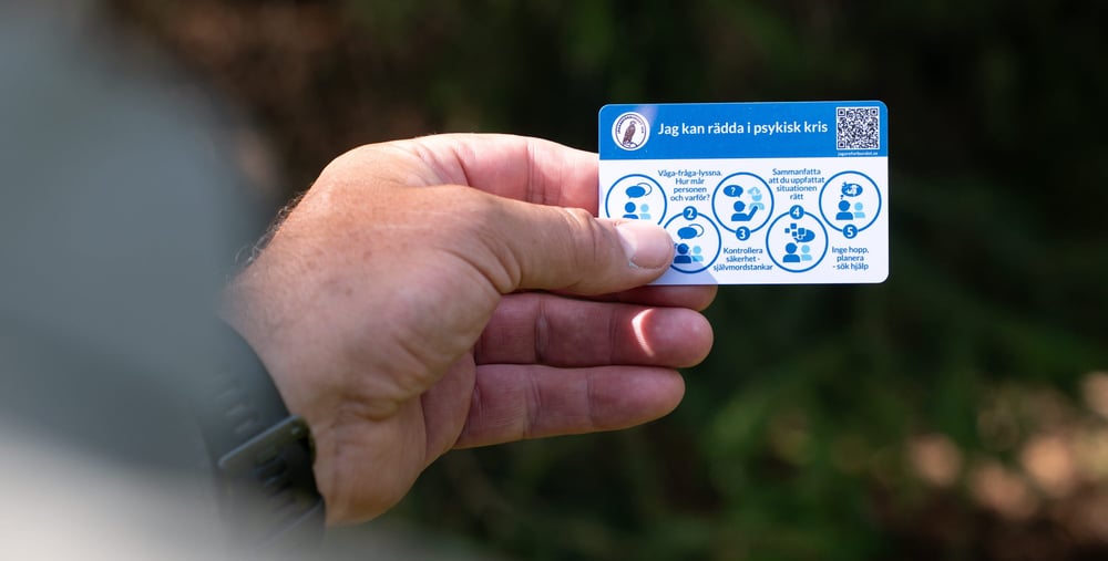 En hand håller ett kort med rubriken "Jag kan rädda i psykisk kris" och fem steg för att hjälpa någon i kris, inklusive att våga fråga, kontrollera säkerhet och söka hjälp.