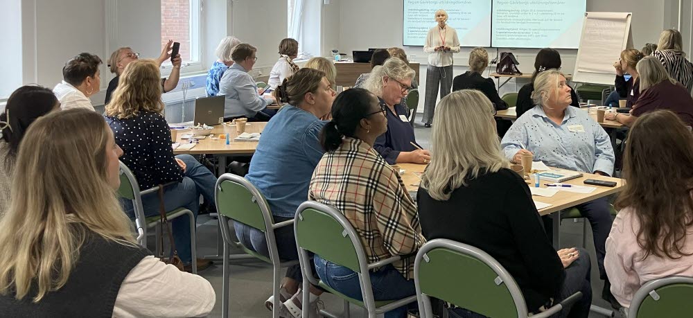 Deltagare sitter vid bord i ett konferensrum och lyssnar på en presentation som visas på storbildsskärm.