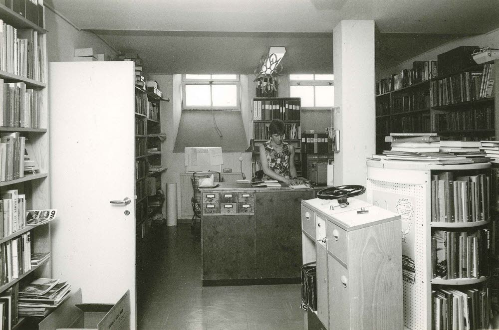 Svartvit bild av ett äldre bibliotek där en kvinna arbetar bakom en trädisk med kortkatalog. Rummet är fullt av bokhyllor, pärmar och dokument i ett trångt men organiserat utrymme.