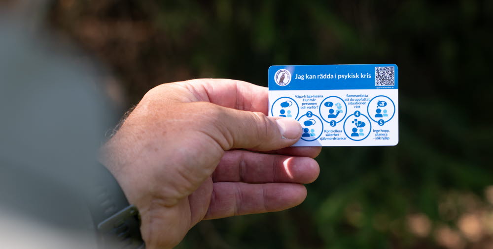 En hand håller ett kort med rubriken "Jag kan rädda i psykisk kris" och fem steg för att hjälpa någon i kris, inklusive att våga fråga, kontrollera säkerhet och söka hjälp.