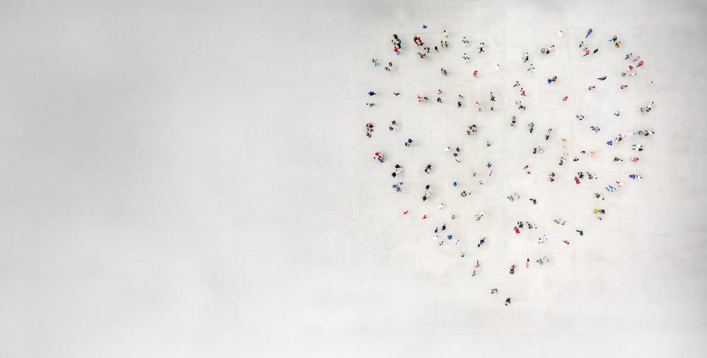 Människor står utspridda så att de tillsammans bildar formen av ett hjärta på en stor öppen yta, sedd uppifrån.