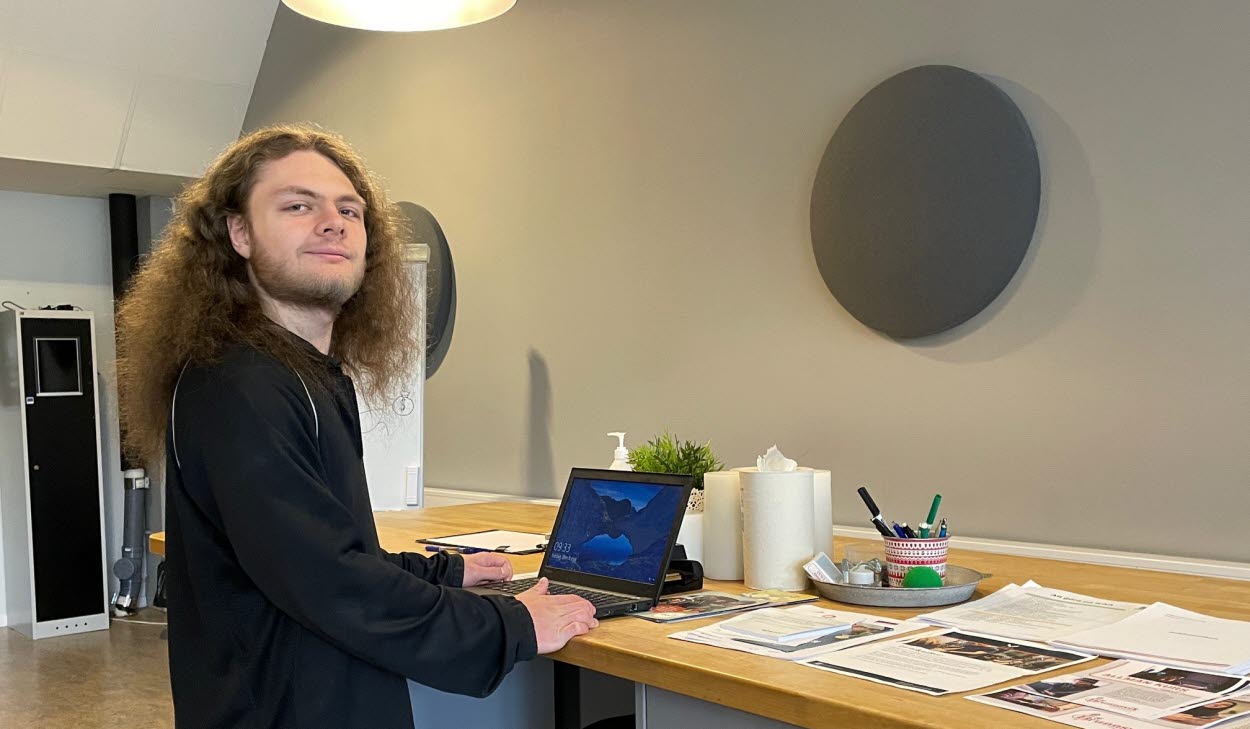 Person vid en dator ståendes vid en arbetsbänk med papper liggandes på sidan.