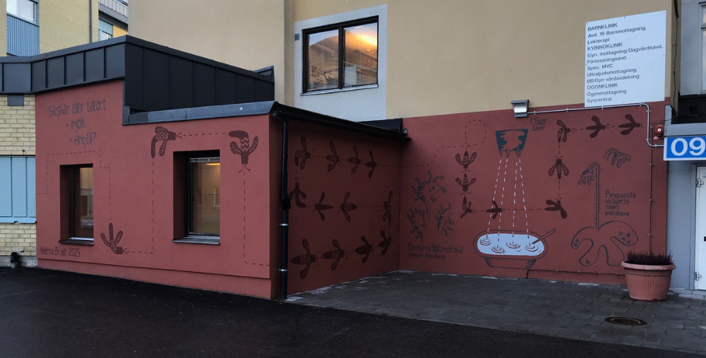 Väggmålning med växtmotiv och text pryder en husfasad vid en entré.