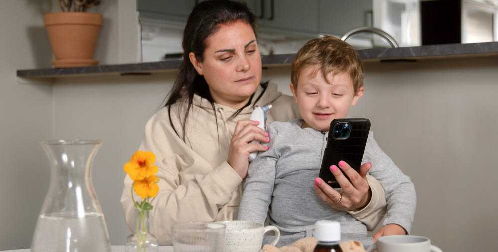Mamma med barn i knä håller i telefon och termometer